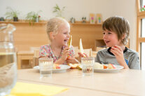 Zwei Kinder sitzen hinter Tellern und Gläsern an Tisch und halten Birnenspalte in der Hand